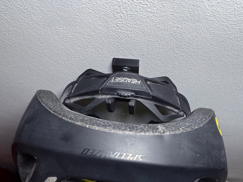 Puede incluir: Un casco de bicicleta Specialized negro con un soporte de casco negro fijado a una pared blanca. El soporte de casco tiene la palabra "HEADSET" impresa.
