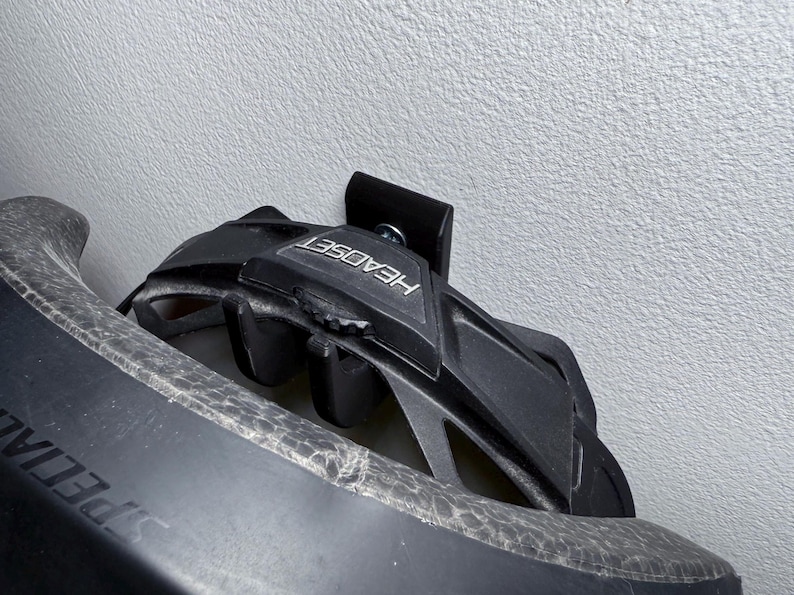 Puede incluir: Un casco de bicicleta negro con la palabra "HEADSET" impresa en el lateral, colgado de un soporte de pared negro.