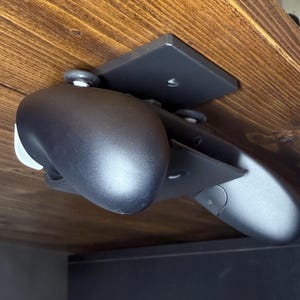 May include: A black under-desk computer mount with a rounded, ergonomic design. The mount is attached to a wooden surface with a square bracket and small wheels for smooth movement. The design is sleek and modern.