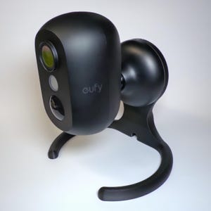 May include: A black security camera with the brand name "eufy" printed on the side. The camera has a rounded shape and is mounted on a black stand. The camera lens is visible, and the overall design is modern and sleek.