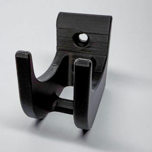Puede incluir: Un soporte de pared negro impreso en 3D con dos ganchos. El soporte tiene un agujero para montaje.