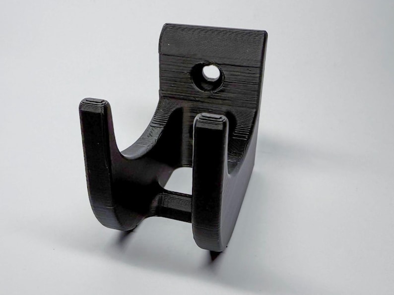 Puede incluir: Un soporte de pared negro impreso en 3D con dos ganchos. El soporte tiene un agujero para montaje.