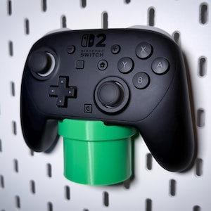 Peut inclure: Une manette Nintendo Switch Pro noire montée sur un support vert en forme de tuyau. La manette a des accents gris et des étiquettes de boutons. Le support est fixé à une planche perforée blanche avec des trous espacés.