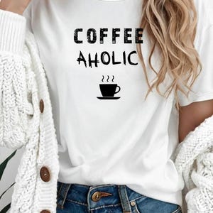 Może przedstawiać: Biała koszulka z czarnym napisem "COFFEE AHOLIC" i czarnym rysunkiem filiżanki kawy. Koszulka jest połączona z niebieskimi dżinsami i białym kardiganem.