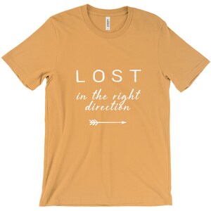 May include: Mustard-colored t-shirt with the text "LOST in the right direction" in white, along with a white arrow pointing right. The shirt is made of a soft material and has short sleeves.