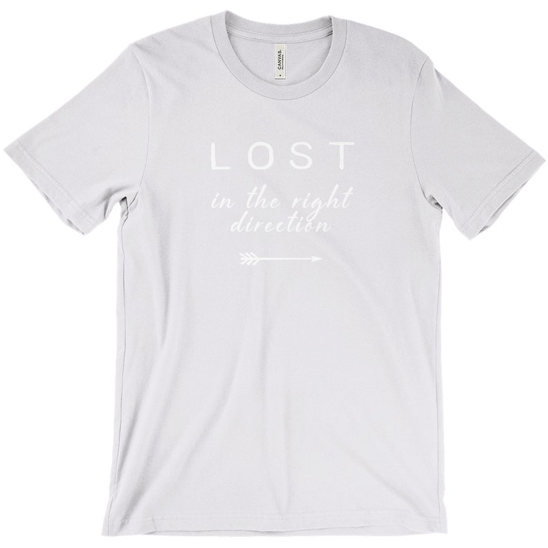 May include: A light gray t-shirt with the words "LOST in the right direction" printed in white. An arrow graphic is below the text. The shirt has a crew neck and short sleeves.