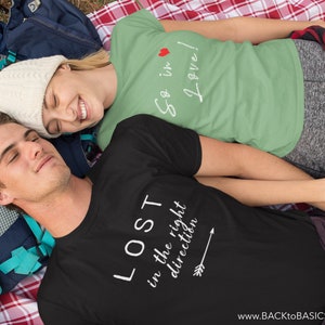 May include: Two people wearing t-shirts with text are lying on a blanket. One shirt is black with the text "LOST in the right direction" and an arrow. The other shirt is green with the text "So in Love!" and a heart. A backpack is visible.