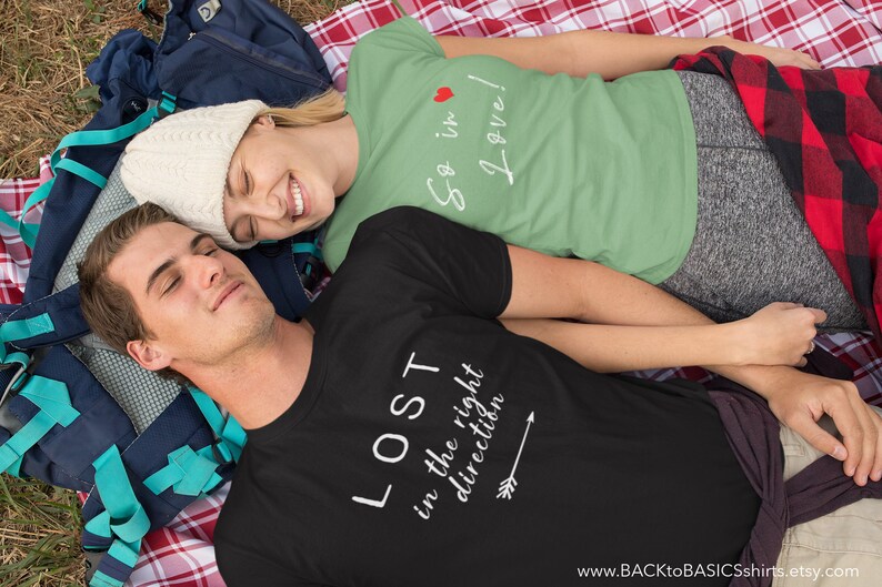 May include: Two people wearing t-shirts with text are lying on a blanket. One shirt is black with the text "LOST in the right direction" and an arrow. The other shirt is green with the text "So in Love!" and a heart. A backpack is visible.