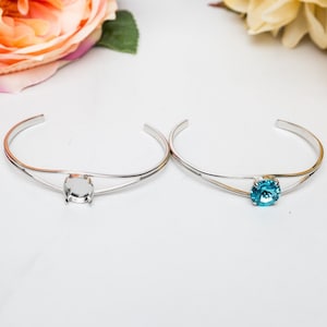 May include: Two silver bangle bracelets with a single round bezel setting. One bracelet has a clear bezel setting and the other has a turquoise blue stone.