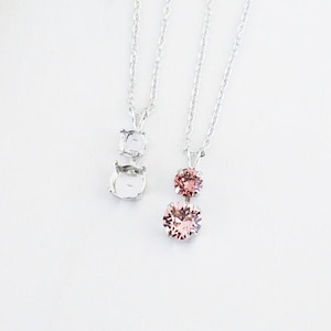 May include: Two silver necklaces with delicate chains. One necklace features a single pink crystal pendant, while the other has a double crystal pendant.