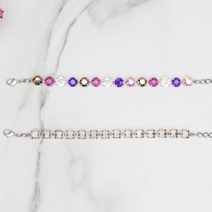 May include: Two silver bracelets with a chain link design. The top bracelet has a gradient of purple, pink, and white round crystals. The bottom bracelet has clear round crystals.
