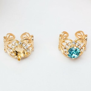May include: Two gold ear cuffs with intricate designs. One cuff features a clear rhinestone center, while the other has a blue rhinestone center.