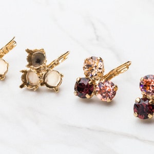 May include: Three pairs of gold-tone earring settings with three round clear crystal settings. The earrings are shown on a white marble surface.