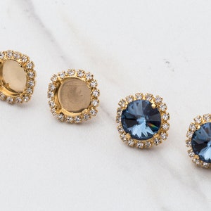 May include: Four gold-toned earring settings with clear rhinestone accents. Two settings are empty and two settings feature a dark blue gemstone.