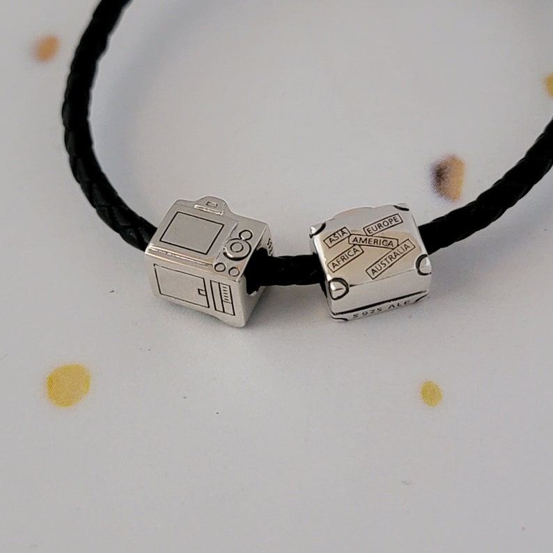 May include: A silver charm bracelet with two charms. One charm is a camera and the other charm is a cube with the words "Asia", "Europe", "America", "Africa", "Australia" and "925 ALE" on it.