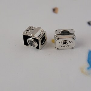 May include: Two silver charm beads for a bracelet. One bead is a black and silver camera with a clear crystal lens. The other bead is a silver suitcase with the text "I &hearts; TO TRAVEL" on it.