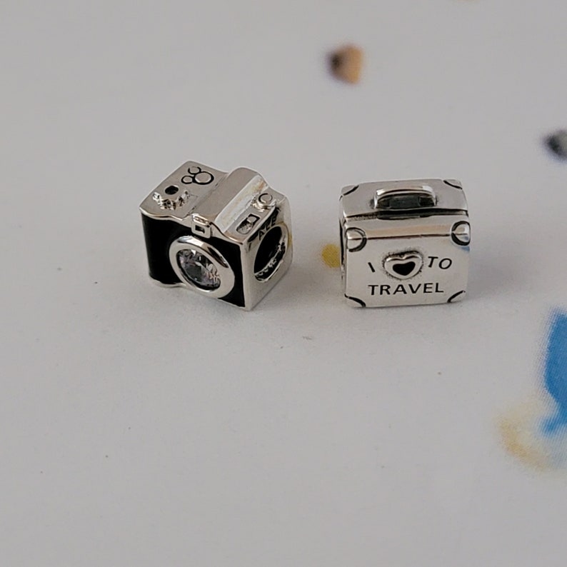 May include: Two silver charm beads for a bracelet. One bead is a black and silver camera with a clear crystal lens. The other bead is a silver suitcase with the text "I &hearts; TO TRAVEL" on it.