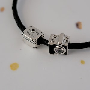 May include: Two silver charm bracelets with a black leather cord. One charm is a silver suitcase with the text "I &hearts; TO TRAVEL" and the other charm is a silver camera with a clear crystal.