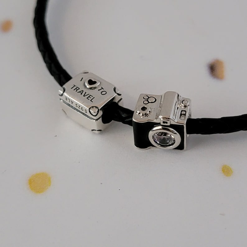 May include: Two silver charm bracelets with a black leather cord. One charm is a silver suitcase with the text "I &hearts; TO TRAVEL" and the other charm is a silver camera with a clear crystal.