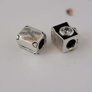 May include: Two silver charm beads for a bracelet. One bead is a suitcase with the words "Asia", "America", "Europe", "Africa", and "Australia" on it. The other bead is a camera with a black face and a clear crystal.