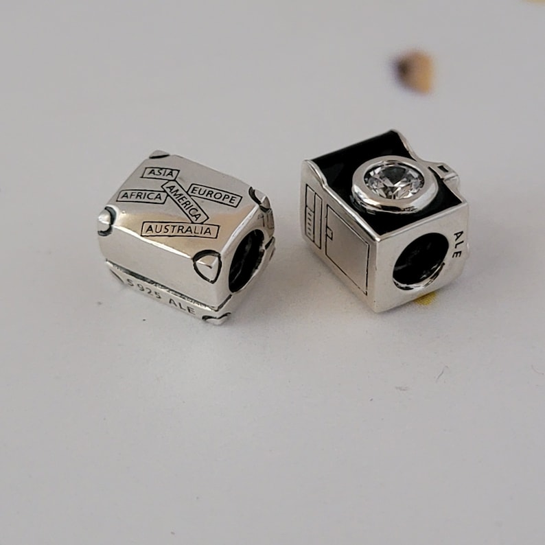 May include: Two silver charm beads for a bracelet. One bead is a suitcase with the words "Asia", "America", "Europe", "Africa", and "Australia" on it. The other bead is a camera with a black face and a clear crystal.