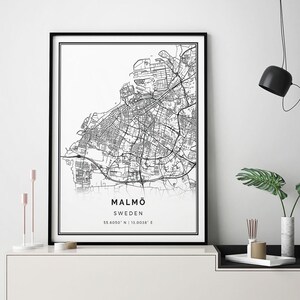 May include: Black and white line art print of a map of Malmö, Sweden. The print is framed in a black frame and is hanging on a white wall above a white console table with a black top. The console table has a black metal frame and is sitting on a white floor.