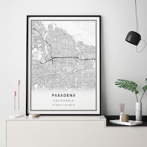 May include: Black and white map print of Pasadena, California in a black frame. The map is on a white shelf with a black and white side table. The shelf also has a vase with green plants, a small white tray with incense sticks, and a small brown object.