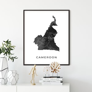 May include: Black and white print of a map of Cameroon with the word "Cameroon" printed below the map. The print is framed in a black frame and is hanging on a white wall.
