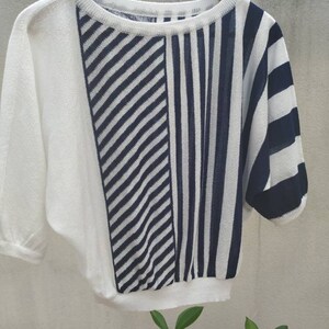 Könnte beinhalten: Ein Strickpullover mit Color-Block-Design, bestehend aus einer einfarbig weißen linken Seite und einer rechten Seite, die in diagonale und vertikale marineblaue und weiße Streifen unterteilt ist. Das Kleidungsstück hat einen Rundhalsausschnitt.