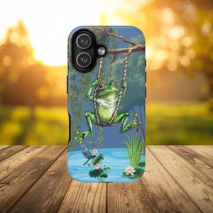 May include: A phone case featuring a green frog swinging on a rope swing. The background depicts a pond with lily pads, a dragonfly, and a sunset. The case has a blue and green color scheme.