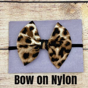 May include: A brown and black leopard print bow on a black nylon headband. The bow is tied in a knot and the headband is on a purple background. The text "BOW on NYLON" is below the bow.