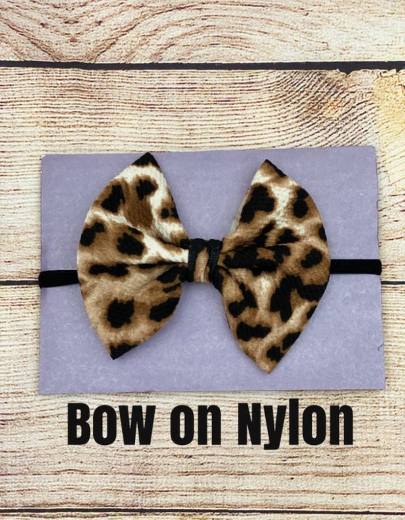 May include: A brown and black leopard print bow on a black nylon headband. The bow is tied in a knot and the headband is on a purple background. The text "BOW on NYLON" is below the bow.