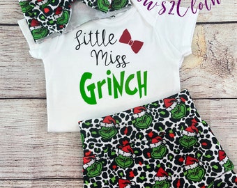 May include: A white onesie with the text "little miss Grinch" and a red bow. A green and white leopard print headband with a Grinch face pattern. A green and white leopard print diaper cover with a Grinch face pattern.