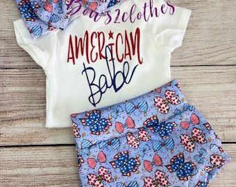 Könnte beinhalten: Ein weißer Baby-Strampler mit dem Text "AMERICAN Babe" in roten und blauen Buchstaben. Der Strampler wird mit blau-weiß gemusterten Shorts mit roten, weißen und blauen Schleifen kombiniert. Ein passendes Stirnband mit dem gleichen Muster ist im Lieferumfang enthalten.
