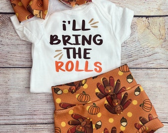 May include: Thanksgiving baby outfit set featuring a white onesie with the text "I'll Bring The Rolls", paired with matching orange turkey and pumpkin print shorts and a large bow. The bow and shorts have a repeating pattern of turkeys, pumpkins, and acorns.
