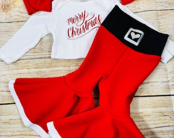 May include: Red and white Christmas outfit for a baby, featuring a white long-sleeved bodysuit with the text "merry Christmas" in red, red bell bottom pants with a black waistband and a silver heart on the front, and a red headband with a bow.
