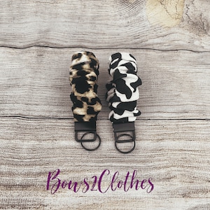 May include: Two fabric keychains with black metal rings. The keychain on the left is brown and black with a leopard print. The keychain on the right is black and white with a cow print. The text "Bows2Clothes" is printed in purple below the keychains.