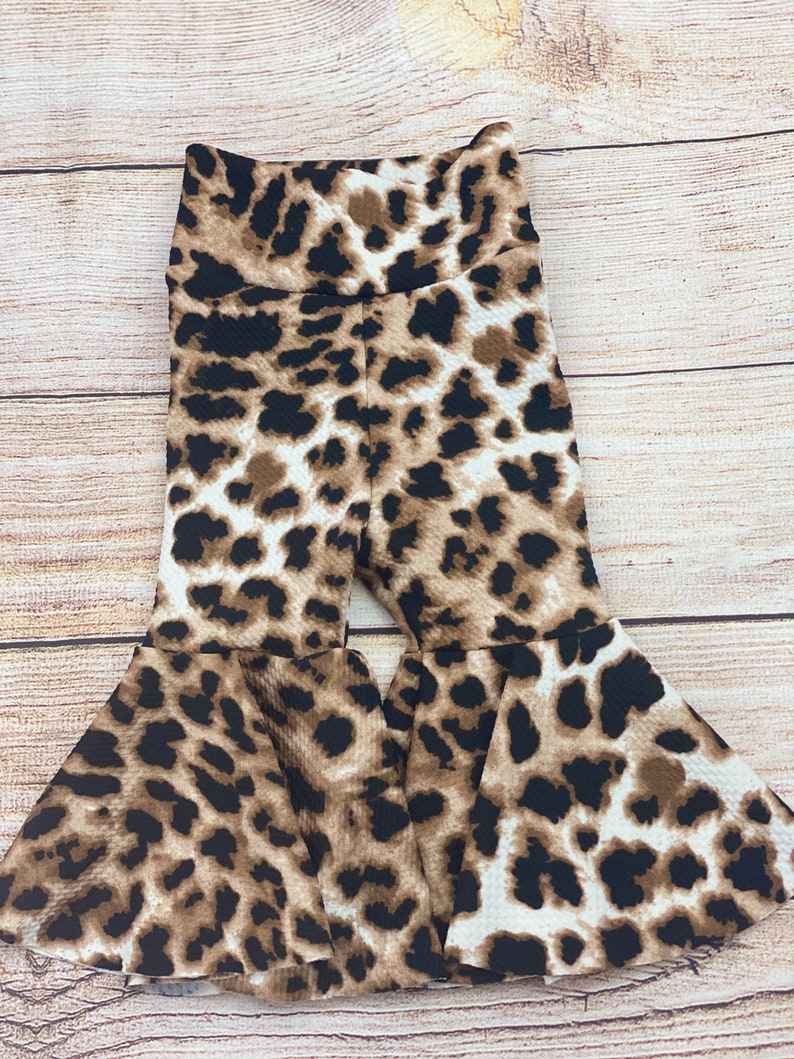 May include: Brown, black, and white leopard print bell bottom pants for a child. The pants have a wide leg opening and a high waist.