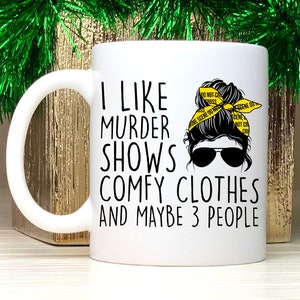 May include: White ceramic mug with a black and white graphic of a woman with a messy bun wearing a yellow "crime scene do not cross" headband. The text on the mug reads "I like murder shows comfy clothes and maybe 3 people."