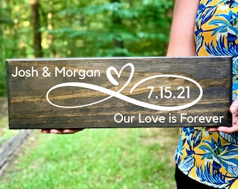 May include: A rectangular wooden sign with the names "Josh & Morgan" and the date "7.15.21" in white script. The sign also features a heart and infinity symbol design, with the phrase "Our Love is Forever" below.