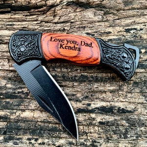 May include: A folding pocket knife with a black blade and a wooden handle. The handle has a wood grain pattern and is engraved with the words "Love you, Dad. Kendra". The knife has decorative black metal accents.