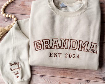 May include: A beige sweatshirt with the word "GRANDMA" embroidered in brown, along with "EST 2024." The left sleeve features embroidered names and a heart. The sweatshirt is displayed on a wooden surface with decorative beads.