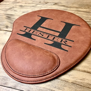 May include: Brown leather mouse pad with a personalized monogram. The letter "H" is centered with the name "HESTER" written below it.