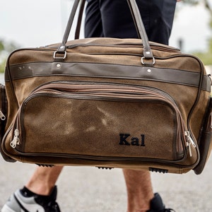 May include: Brown and dark brown duffel bag with multiple zippered compartments and handles. The bag has the name "Kal" embroidered on the front. The bag is carried by a person walking on a paved surface.