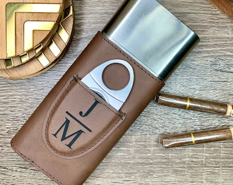 Könnte beinhalten: Ein braunes Leder-Zigarrenetui mit einem silbernen Metalleinsatz und einem integrierten Zigarrenschneider. Das Etui hat eine Tasche für den Schneider und ist mit den Initialen "J/M" personalisiert.