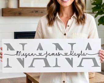 May include: A white wooden sign with gray lettering that reads "NANA" above and below the names "Megan", "James", and "Kayla" in a cursive font. The names are connected by a thin black line with heart symbols. The sign is held by a person.