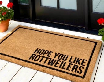 May include: Rectangular doormat made of natural coir fiber with a black border. The text "HOPE YOU LIKE ROTTWEILERS" is printed in black across the mat. The mat is placed on a white wooden porch, with potted red geraniums and a black door in the background.