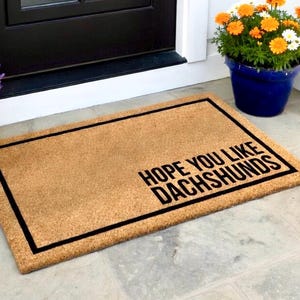 May include: A rectangular doormat made of natural coir fibre, featuring a black border and the text "HOPE YOU LIKE DACHSHUNDS" in bold black letters. The mat is placed in front of a black door, with potted flowers nearby.
