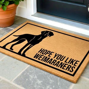 May include: A rectangular doormat with a black border and a tan background. The mat features a black silhouette of a dog and the text "HOPE YOU LIKE WEIMARANERS" in black. The mat is placed in front of a black door.