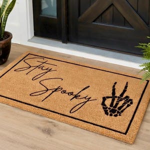 May include: A rectangular doormat made of natural coir fiber. The mat features the phrase "Stay Spooky" in a black script font, along with a black skeleton hand making a peace sign. The mat is framed with a black border.